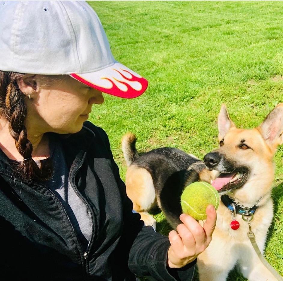 Susan Rosetti of Soozy Pooch crouching in a sunny park, offering a tennis ball to her attentive German shepherd, G